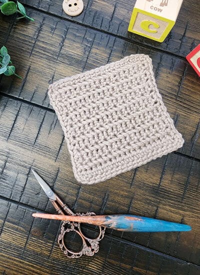 A beige crochet bonding square made using the herringbone single crochet stitch, displayed with a wooden crochet hook, ornate scissors, and baby blocks on a dark wooden surface.