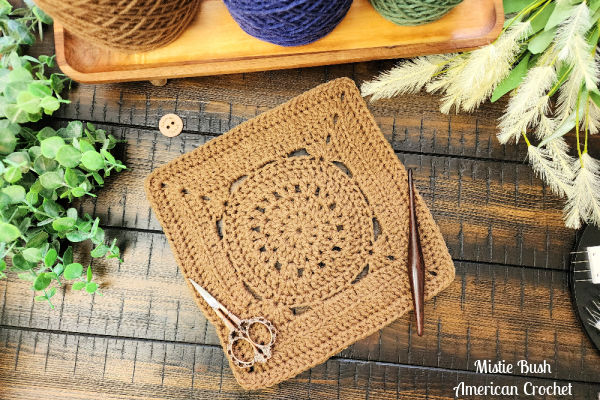 The Pilot Square crochet pattern laid flat with skeins of yarn, a crochet hook, and decorative elements, creating a cozy crafting scene.