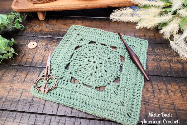 Stable Square crochet block shown flat on a wooden table with greenery and yarn, highlighting stitch texture and openwork spacing.