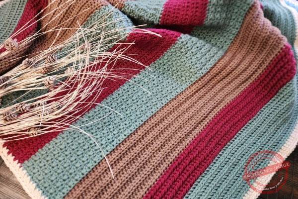 Handmade crochet lapghan featuring horizontal stripes in earthy tones of green, burgundy, and brown, displayed on a wooden surface with a rustic bundle of dried wheat and pinecones resting on top. The textured stitches and cozy palette evoke a sense of warmth and seasonal comfort.